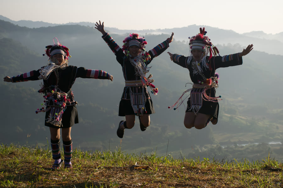 Tres personas vestidas con trajes tradicionales de la etnia Akha saltando alegremente en una colina de pasto, con un paisaje montañoso y neblina al fondo.