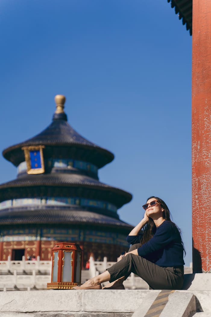 Mujer con lentes de sol sentada frente al icónico Templo del Cielo en Pekín difuminado al fondo bajo un cielo azul