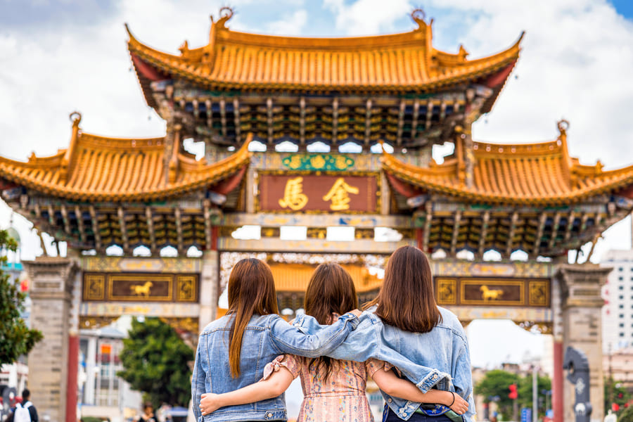 Tres mujeres jóvenes de espaldas y abrazadas por los hombros contemplan una gran puerta de arco tradicional china con techos de tejas doradas y caracteres tradicionales
